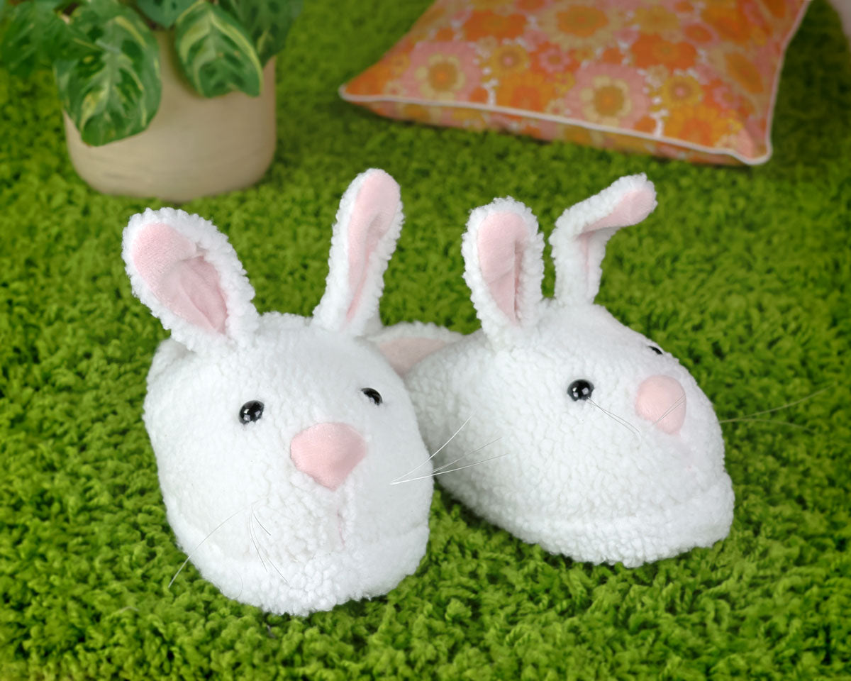 Pair of white bunny slippers with pink noses on a green carpet with a plant and a pillow in the background