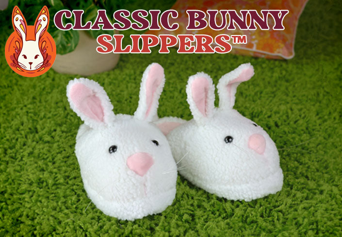 Pair of white bunny slippers with pink ears and noses on a green carpet, featuring the brand 'Classic Bunny Slippers'.