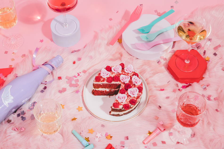 Cake, bottle of wine, and some glasses filled with drinks, surrounded by confetti with a pink background.