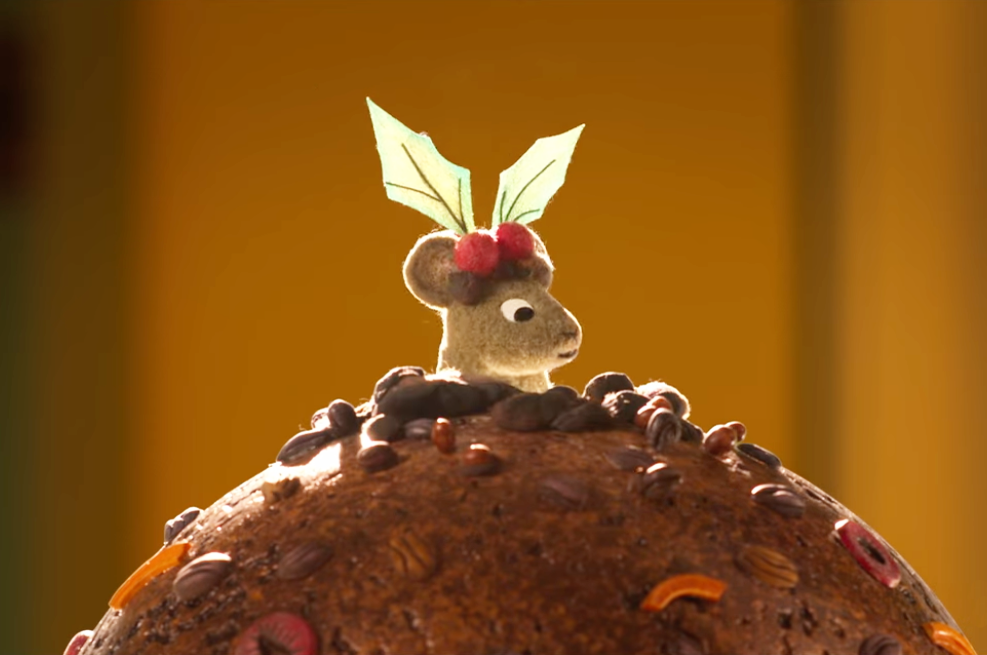A felt mouse looks out from behind a Christmas pudding, with holly branches on its head