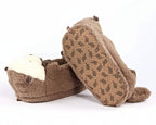 A pair of plush brown slippers shaped like otters, with one slipper showing the white faux-fur face and the other showing the patterned brown sole, set against a plain white background.