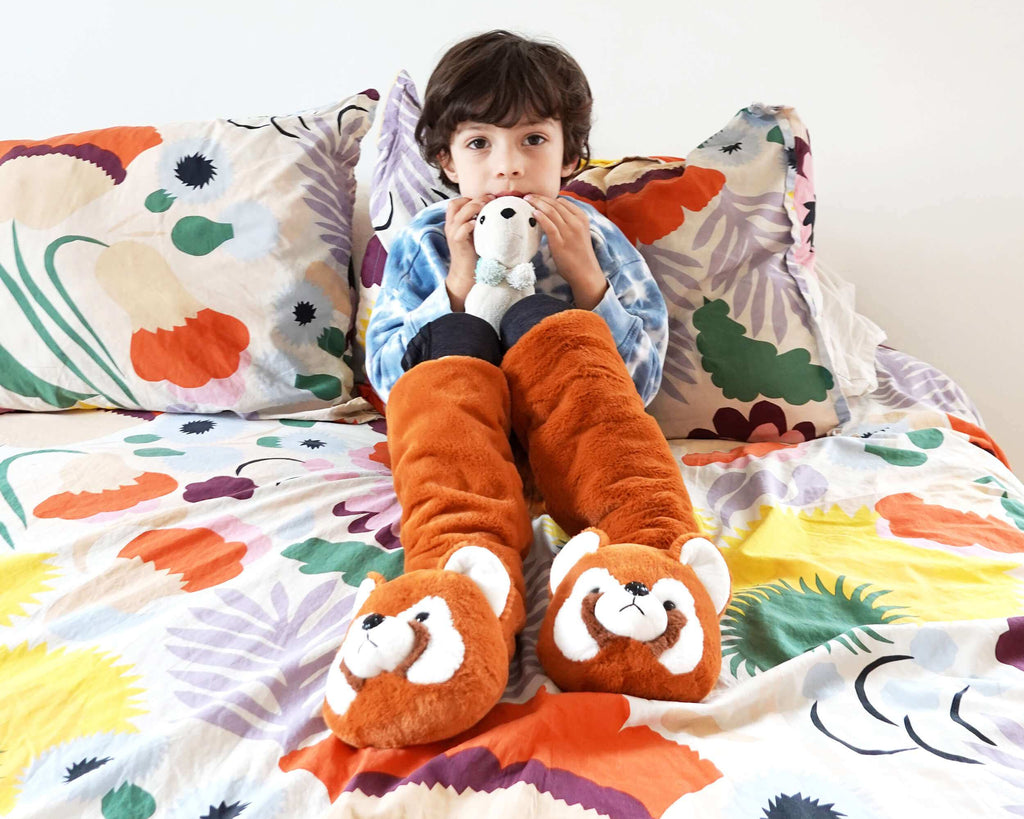 A child sitting on a bed wearing plush orange and white animal slippers with a blue patterned shirt, holding a small stuffed animal. The bed is covered in a colorful floral-patterned duvet against a plain white wall.