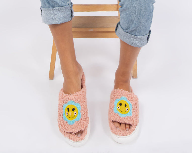 A woman sits on a wooden stool against a white studio background, wearing a white sleeveless crop top, light blue ripped denim shorts, and pink fuzzy slide sandals with yellow smiley faces on the front.