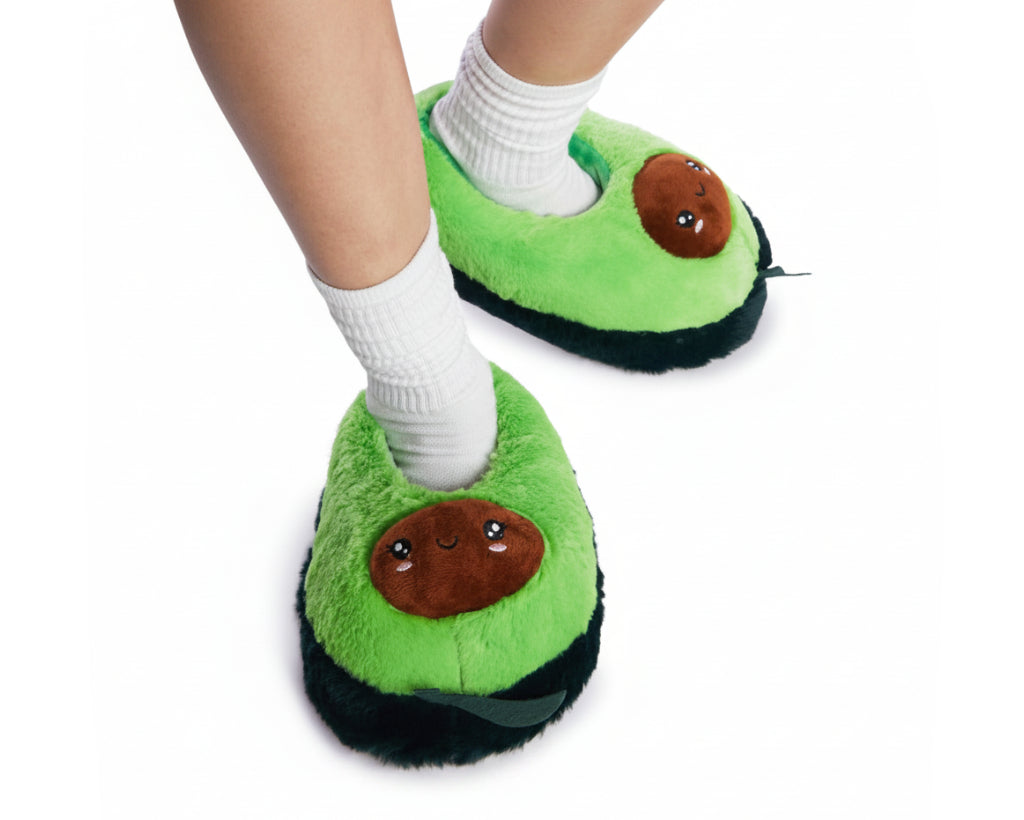 Green avocado-themed plush slippers with brown seed details, shown on a person's feet with white socks against a light gray background.