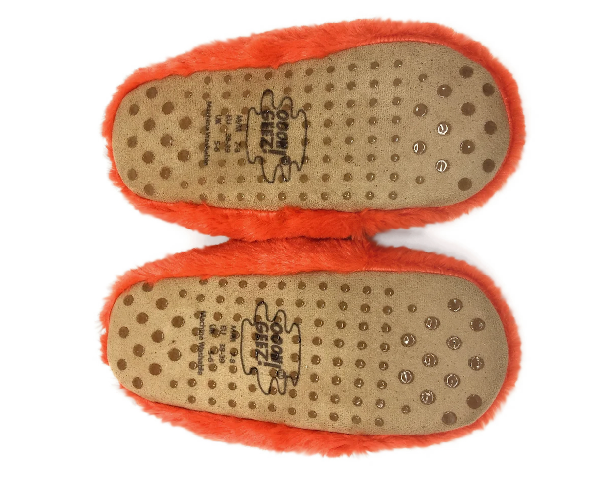 Bottom view of a pair of fluffy orange slippers showing the tan non-slip soles with printed brand markings and rubber grip dots.
