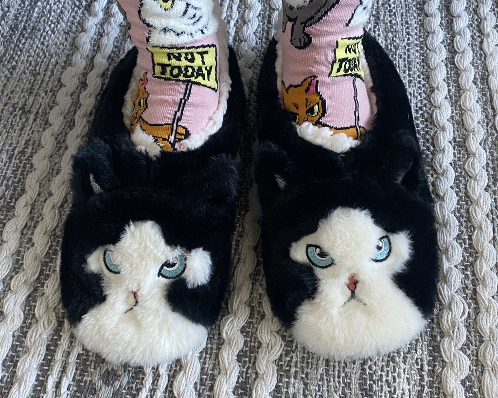 Person's feet wearing pink cat socks and black and white cat slippers standing on a woven rug