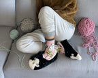 Person sitting on a couch with yarn and knitting needles, wearing a brown sweater and white pants.